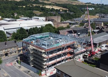 Aerial view of a multi‑storey steel‑frame building under construction with scaffolding and a pink crane, surrounded by roads, warehouses and green hills.