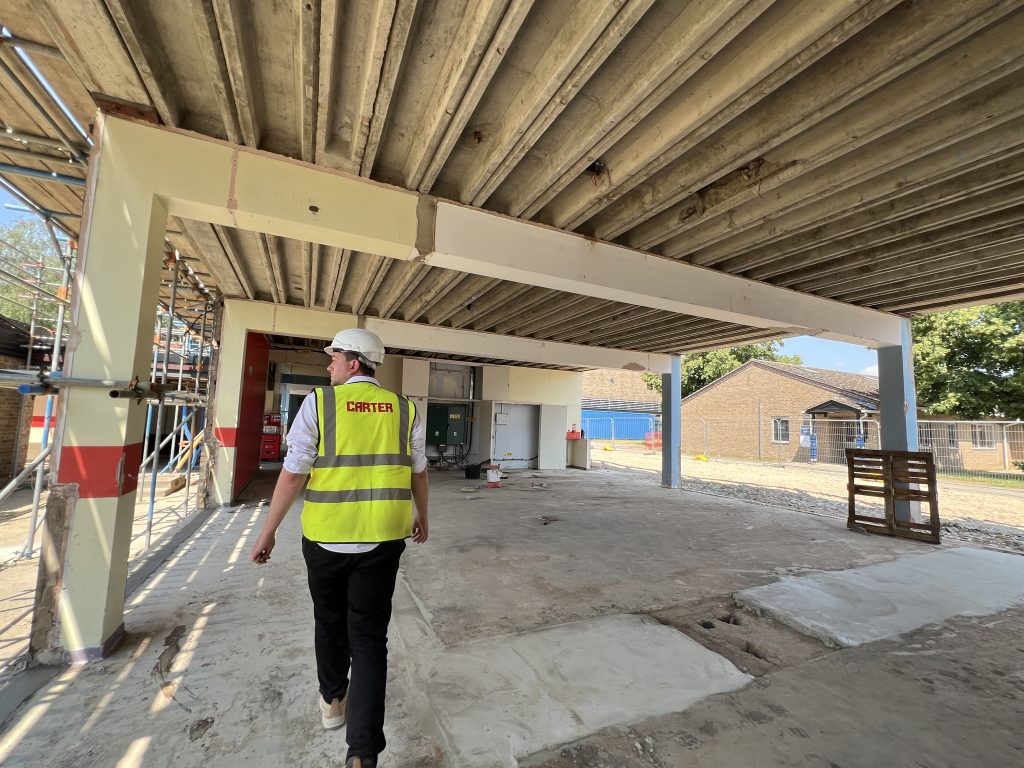A construction worker in a high‑visibility vest and hard hat walks through the stripped‑back Oswald Building at Abbey College Ramsey during refurbishment works.