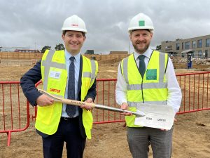 Two construction professionals wearing white hard hats and yellow high‑visibility vests stand on a construction site holding a ceremonial shovel, with red safety barriers and an NHS building visible in the background.