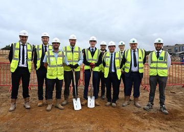 A group of nine people wearing white hard hats and high‑visibility vests stand together on a construction site during a groundbreaking ceremony, with two representatives holding shovels in front of red safety barriers and hospital buildings in the background.