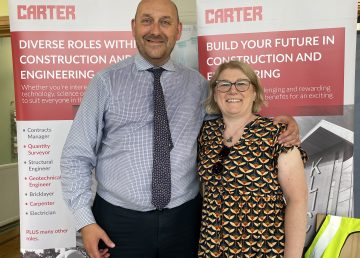 Two R G Carter team members stand at a careers stand with construction and engineering banners behind them, showcasing diverse industry roles. A table in front displays leaflets, coloured pencils and a high‑visibility vest.