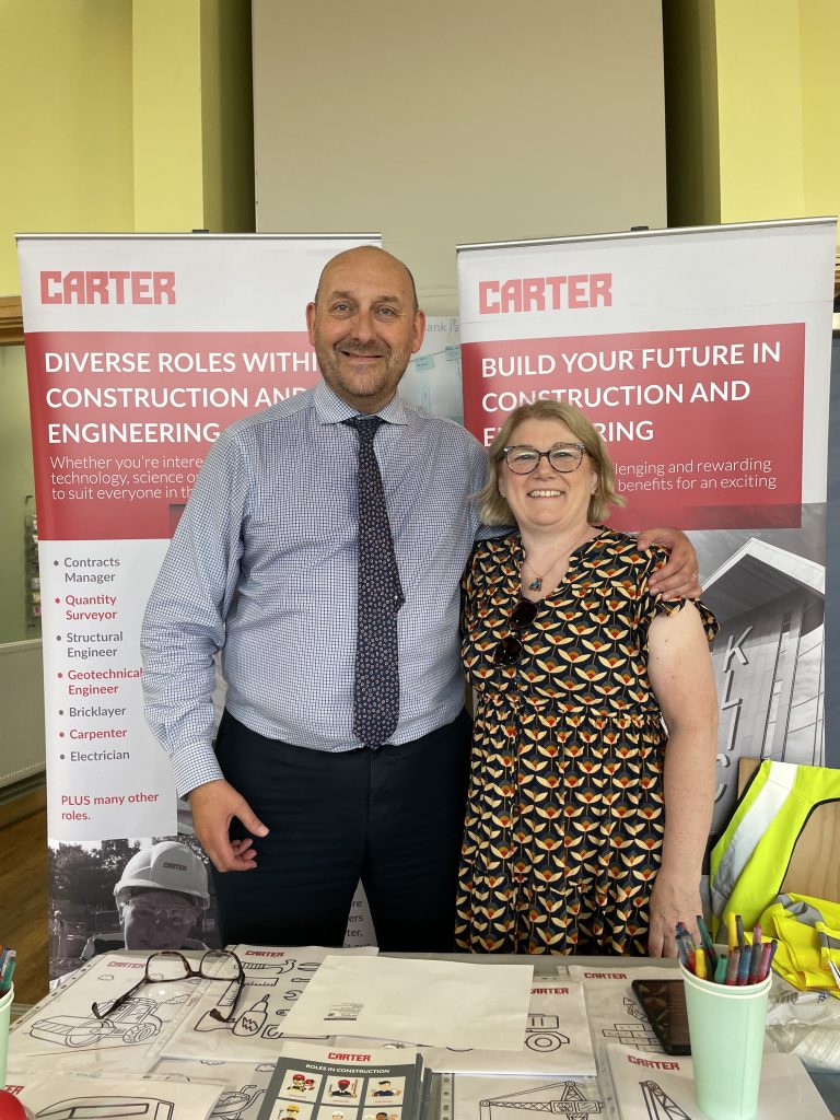 Two R G Carter team members stand at a careers stand with construction and engineering banners behind them, showcasing diverse industry roles. A table in front displays leaflets, coloured pencils and a high‑visibility vest.