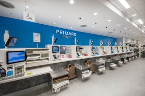 Self‑checkout area inside the refurbished Primark Harlow store, with blue Primark Harlow signage, touchscreen tills and paper bag stations.