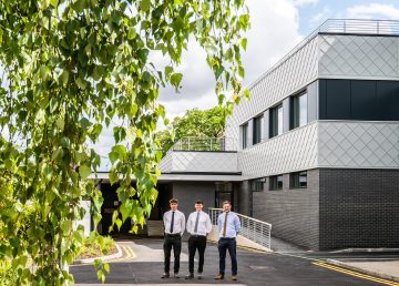 Three members of the project team standing outside the newly completed UEA Edith Cavell Building extension, with dark brickwork, aluminium shingle façades and large upper‑floor windows framed by greenery in the foreground.