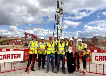 A group of nine representatives from R G Carter, Norfolk County Council, Homefield Primary School, NPS and Roy Sykes Consulting stand together in hard hats and high‑visibility vests at the groundbreaking event for the new Homefield C of E VC Primary School in Bradwell, with construction machinery and red CARTER safety barriers behind them.