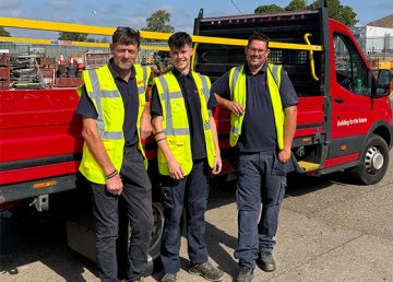 Three operatives in high‑visibility vests stand beside a red R G Carter flatbed truck in the Thetford plant yard, with construction materials and equipment visible in the background.