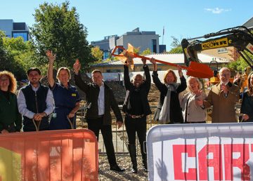 Representatives from R G Carter, Maggie’s and project supporters gather on the construction site of the new Maggie’s Cambridge centre for a ceremonial groundbreaking event, holding a commemorative orange shovel in front of ongoing site works and construction equipment.
